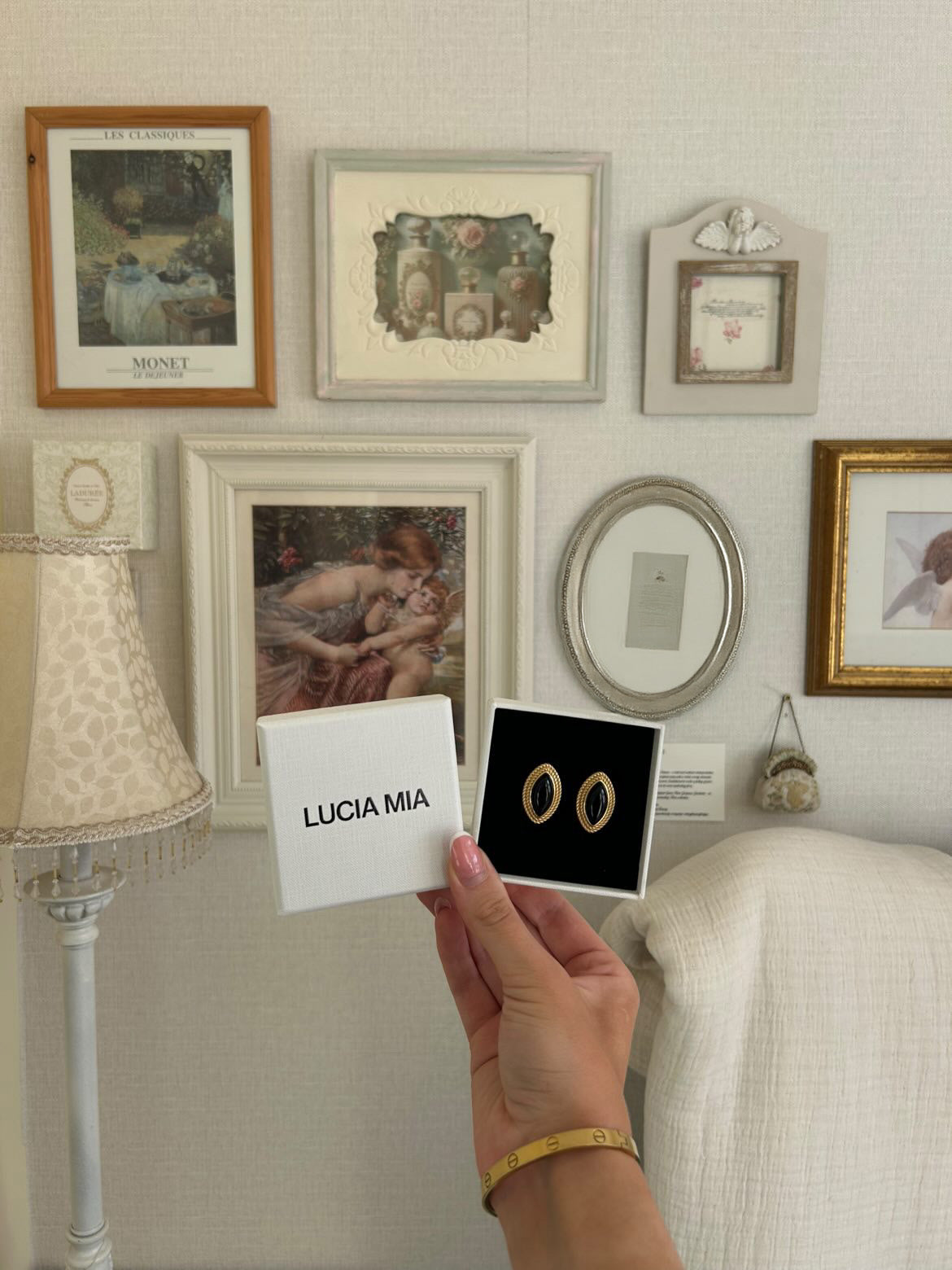 Person holding a box of earrings with 'LUCIA MIA' branding in front of a wall with framed pictures.
